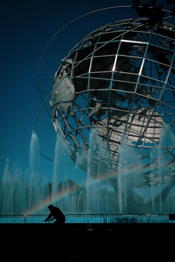 Silhouette of a cyclist near the iconic Unisphere in Flushing Meadows, NYC, with fountains and rainbow.