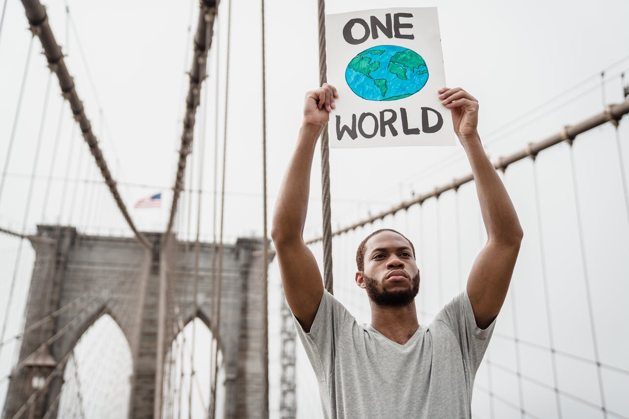A man holds a One World sign on the Brooklyn Bridge, symbolizing unity and activism.